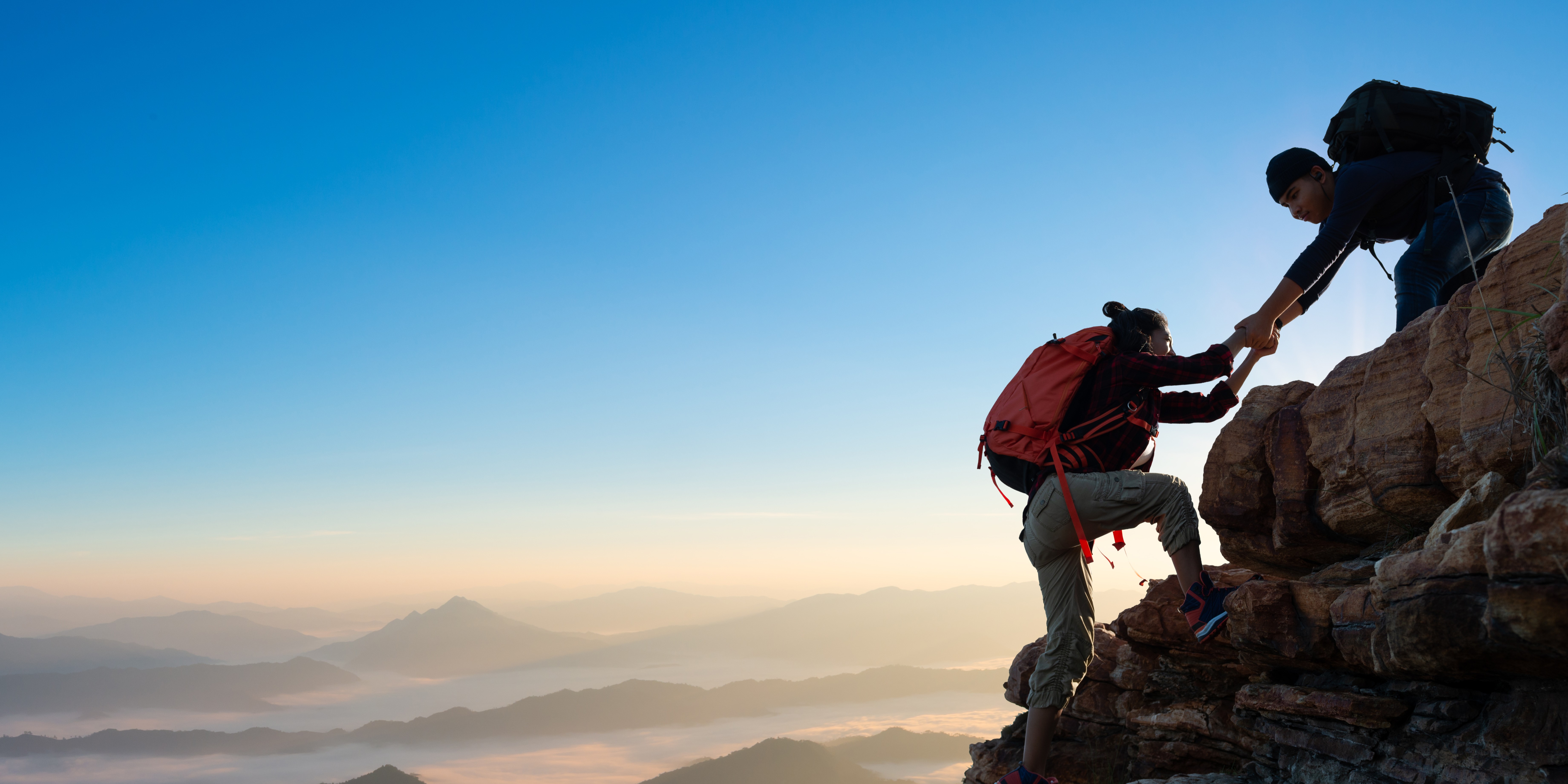 one hiker helping another to the top of a peak